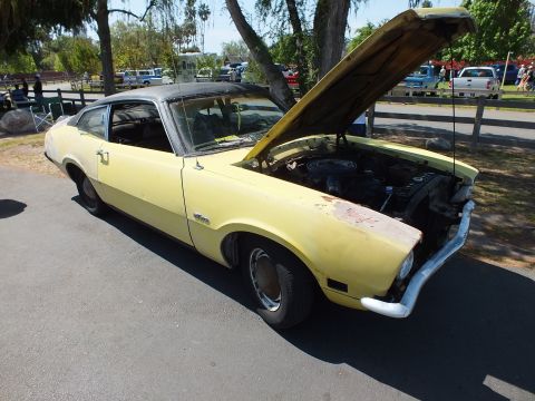 2003 Ford Crown Victoria - yellow Ford Maverick two-door coupe with primer patches and its hood up, parked on pavement near a fence at an outdoor event.
