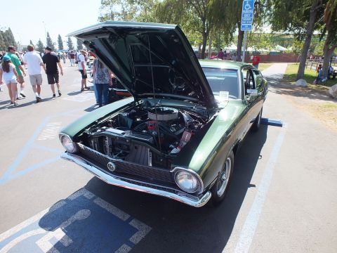 2003 Ford Crown Victoria - dark green Ford Maverick coupe with its hood raised, showing a detailed engine, parked in a handicap stall amid a crowd at a show.