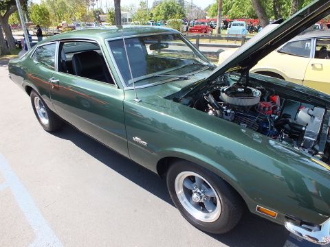 2003 Ford Crown Victoria - Side view of a dark green Ford Maverick Grabber coupe with chrome wheels and its hood raised, parked among show cars on pavement.