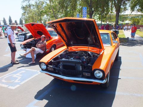 2003 Ford Crown Victoria - orange Ford Maverick two-door coupe with its hood up sits in a parking stall while a seated man and onlookers gather nearby.