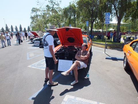2003 Ford Crown Victoria - orange coupe with its hood up and a printed info placard leaning against it, as two men converse at a sunny outdoor car show.