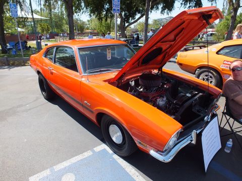 2003 Ford Crown Victoria - orange Ford Maverick Grabber coupe with black steel wheels and a chromed engine visible under the raised hood at a tree-lined show.