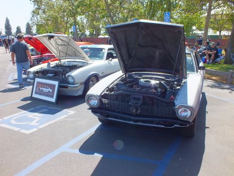 2003 Ford Crown Victoria - Two coupes with raised hoods sit in handicap stalls, a silver-and-black car in front with a framed photo propped beside it at a car show.