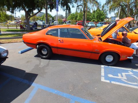 2003 Ford Crown Victoria - Side profile of an orange Ford Maverick two-door coupe with a black side stripe and black steel wheels, parked at an outdoor car show.