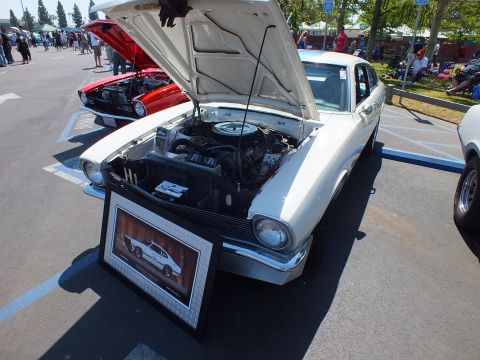 2003 Ford Crown Victoria - white Ford Maverick coupe with its hood up and a framed photo placard leaning against the bumper, parked among show cars.
