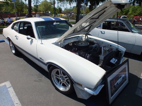 2003 Ford Crown Victoria - white Ford Maverick coupe with a blue roof stripe, polished five-spoke wheels and its hood raised, parked at a tree-lined car show.