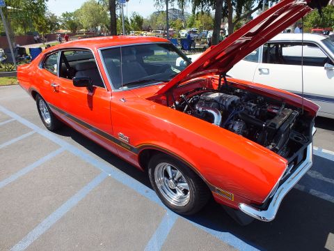 2003 Ford Crown Victoria - red Ford Maverick Grabber coupe with a gold side stripe, polished wheels and its hood raised, parked among other cars at an outdoor show.