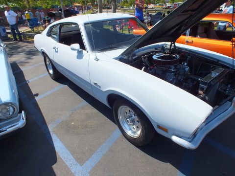 2003 Ford Crown Victoria - white Ford Maverick two-door fastback with chrome wheels sits with its hood raised at a sunny car show.