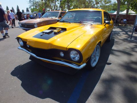 2003 Ford Crown Victoria - bright-yellow Ford Maverick with black hood-scoop stripe and five-spoke wheels sits front-on under sunny skies at a car show.