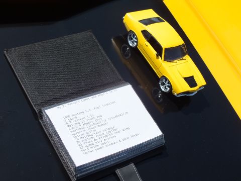 2003 Ford Crown Victoria - open black display book lists a 1990 Mustang 5.0 fuel-injection build alongside a small yellow toy muscle car on a glossy surface.