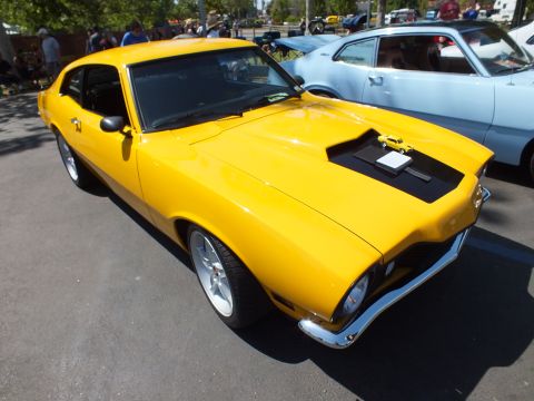 2003 Ford Crown Victoria - yellow Ford Maverick with a black hood scoop and chrome bumper is parked among other cars at a sunny outdoor show.