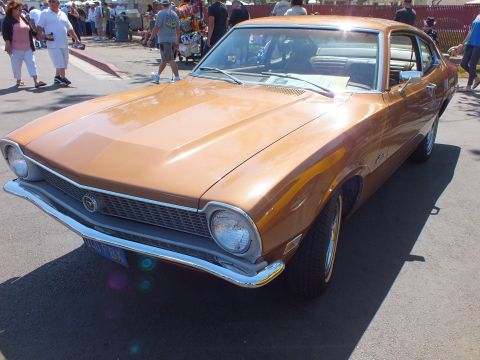 2003 Ford Crown Victoria - copper-brown Ford Maverick two-door fastback sits front-on at a crowded car show, chrome grille and bumper shining in the sun.