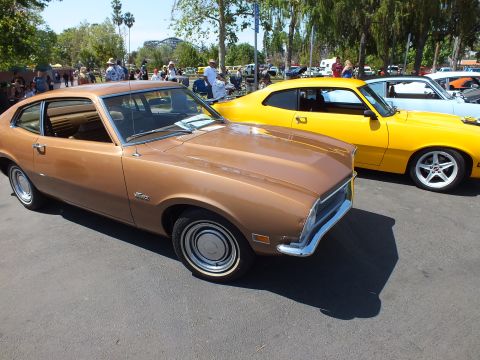 2003 Ford Crown Victoria - brown Ford Maverick with dog-dish hubcaps sits beside a yellow muscle car on a lawn-lined car-show lot full of spectators.