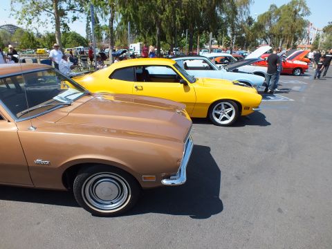 2003 Ford Crown Victoria - row of Ford Mavericks in brown, yellow and blue line up nose-to-tail across a sunny car-show parking lot.