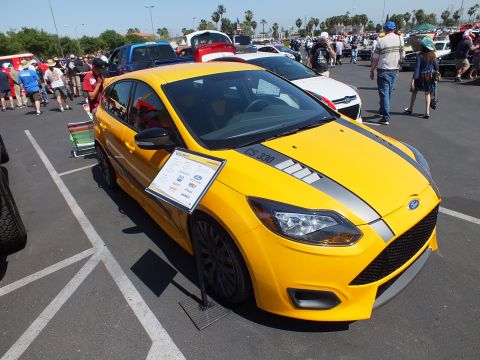 2003 Ford Crown Victoria - yellow Ford Focus hatchback with gray racing stripes and a printed display card is parked at a sunny car show.