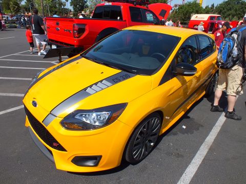 2003 Ford Crown Victoria - yellow Ford Focus ST with gray stripes and black wheels sits beside a red Ford pickup at a busy car show.