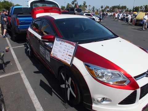 2003 Ford Crown Victoria - red-and-white Ford Focus hatchback with its rear hatch open and a spec placard sits in a crowded car-show lot.