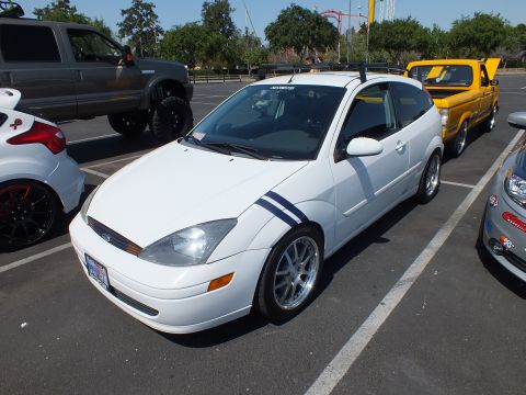 2003 Ford Crown Victoria - white Ford Focus three-door coupe with blue hood-side stripes and chrome wheels parked among other vehicles at an outdoor show.