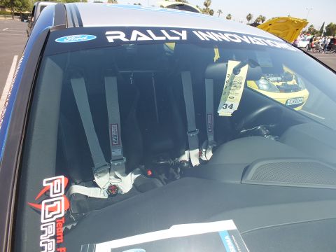 2003 Ford Crown Victoria - Close-up through a windshield showing a Rally Innovations banner and red racing harnesses over bucket seats inside a Ford race car.