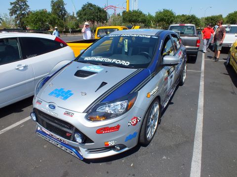 2003 Ford Crown Victoria - Silver Ford Focus ST hatchback rally car with blue hood accents and sponsor decals parked among yellow cars at an outdoor event.