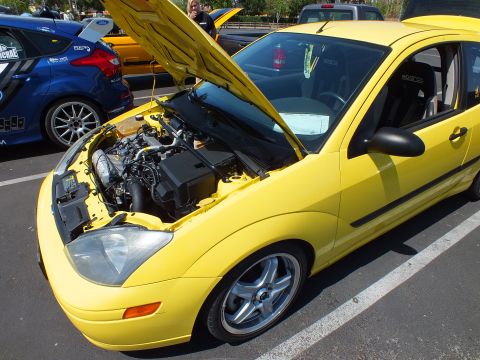 2003 Ford Crown Victoria - yellow Ford Focus three-door coupe with its hood raised showing the engine, parked beside a blue rally car at an outdoor event.