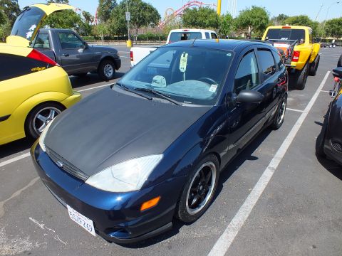 2003 Ford Crown Victoria - dark blue Ford Focus five-door hatchback with black wheels parked beside a yellow truck at an outdoor car gathering.
