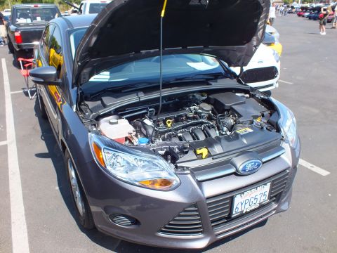 2003 Ford Crown Victoria - gray Ford Focus sedan with its hood raised exposing the engine bay, parked at an outdoor car show with a California plate.