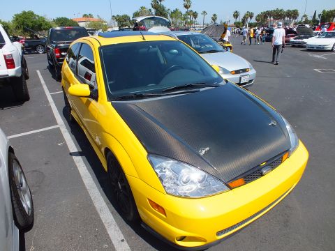2003 Ford Crown Victoria - yellow Ford Focus three-door coupe with a black carbon-look hood and black wheels parked in a busy outdoor car-show lot.