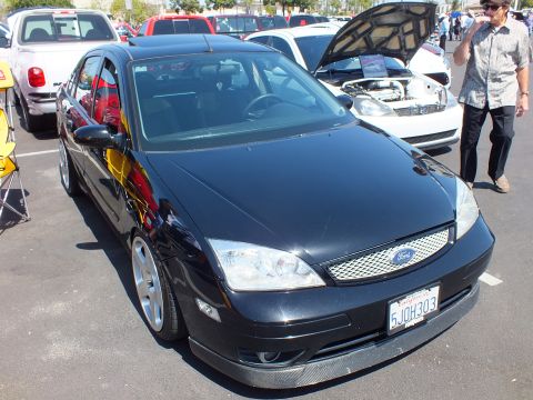 2003 Ford Crown Victoria - glossy black Ford Focus sedan with a chrome mesh grille and silver wheels, plate 5JQH303, at a car meet.