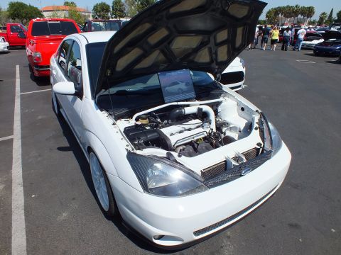 2003 Ford Crown Victoria - white Ford Focus with hood raised displaying the engine bay and a printed info placard at a car show.