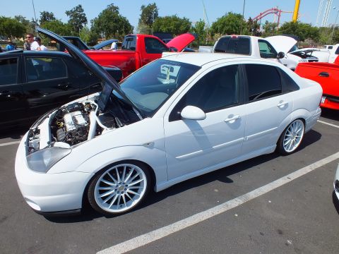 2003 Ford Crown Victoria - white Ford Focus sedan with its hood raised and large silver wheels, parked at a sunny outdoor car meet.