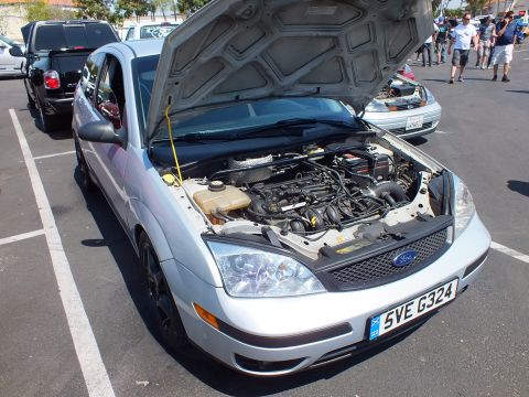 2003 Ford Crown Victoria - silver Ford Focus sits in a parking-lot car show with its hood raised, exposing the engine and a yellow prop rod.