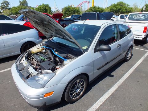 2003 Ford Crown Victoria - silver Ford Focus three-door hatchback has its hood raised in a lot, with an amusement-park coaster in the background.