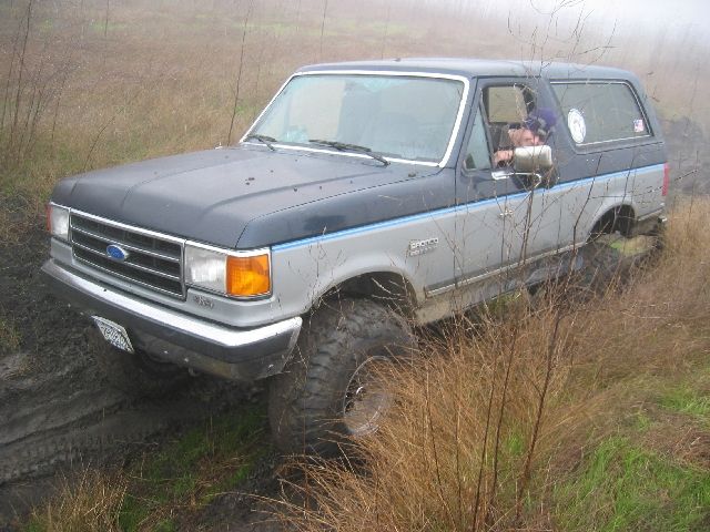 Lifted gray and blue Ford Bronco on mud tires in a foggy, muddy field