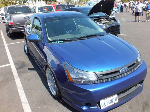 2003 Ford Crown Victoria - lowered blue Ford Focus coupe on chrome lip wheels is parked at a sunny car show, front three-quarter view.