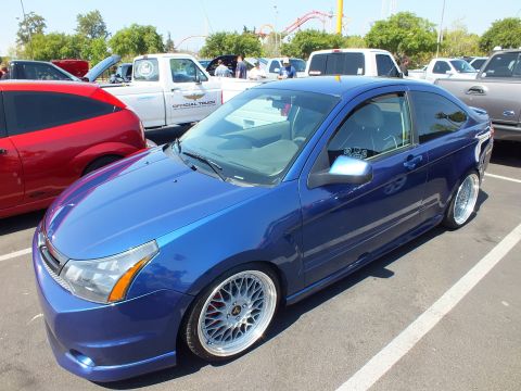 2003 Ford Crown Victoria - lowered blue Ford Focus two-door on silver mesh wheels sits at a car show with a roller coaster in the distance.