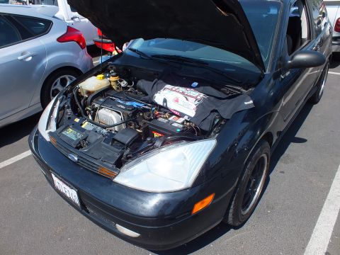 2003 Ford Crown Victoria - black Ford Focus has its hood raised, revealing the engine bay and battery, parked beside other cars in a lot.
