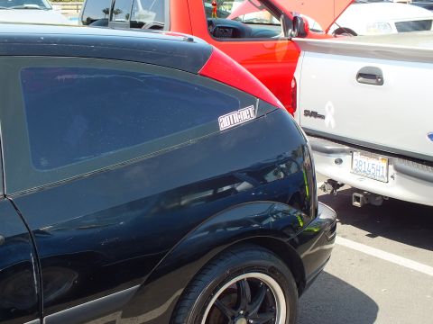 2003 Ford Crown Victoria - black rear quarter panel of a car bears a supermotors.net sticker, parked next to a red and white pickup.