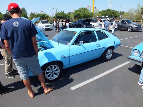 2003 Ford Crown Victoria - light blue Ford Pinto hatchback sits with its hood raised at a sunny outdoor car show as a man in a red cap looks on.