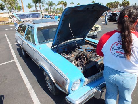 2003 Ford Crown Victoria - weathered blue-and-silver Ford Pinto station wagon shows its engine bay with the hood open while a woman in a raglan tee stands nearby.