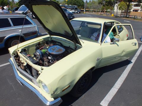 2003 Ford Crown Victoria - pale yellow Ford Pinto with its hood and driver door open reveals the engine bay in a sunlit parking lot car show.