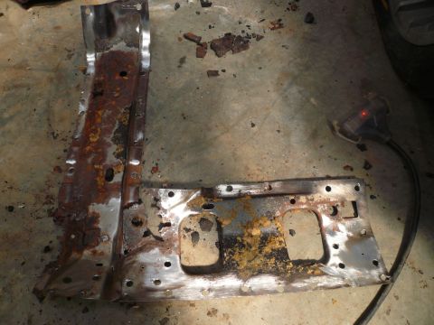 1975 Ford Ranchero - A heavily rusted metal component, possibly from a vehicle's undercarriage, lies on a concrete floor next to a tool.