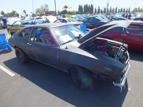 2003 Ford Crown Victoria - flat primer-gray hatchback sits with its hood open over a chromed V8 amid rows of cars at a busy outdoor show.