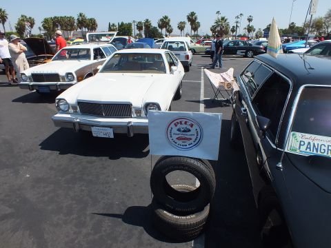 2003 Ford Crown Victoria - white compact coupe displayed at a car show beside a club sign on stacked tires, with another car showing a California license plate.