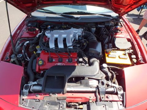 2003 Ford Crown Victoria - Close-up of a red car's engine bay showing a transverse V6 engine with Ford-marked components and a yellow battery.