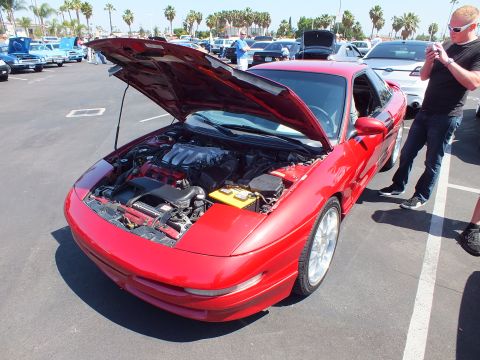 2003 Ford Crown Victoria - red sports coupe with its hood raised at an outdoor car show as a man photographs another vehicle nearby.