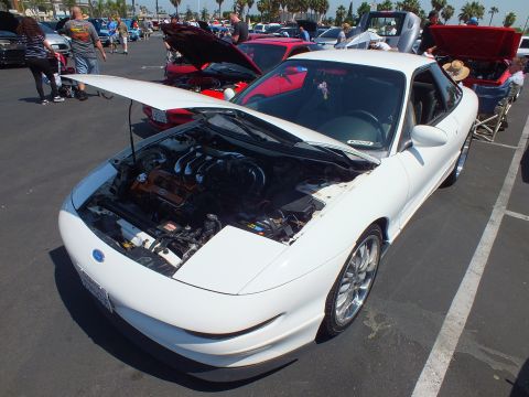 2003 Ford Crown Victoria - white sports coupe with a sunroof and raised hood seen from above at a crowded outdoor car show.