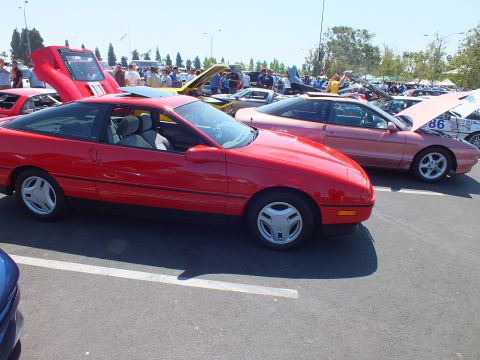 2003 Ford Crown Victoria - red 1980s hatchback coupe shown in profile next to a salmon-colored coupe at a busy outdoor car show.