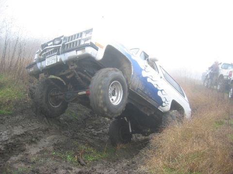 1989 Ford Bronco - This lifted SUV with flame decals navigates a muddy incline, with other vehicles visible in the background.
