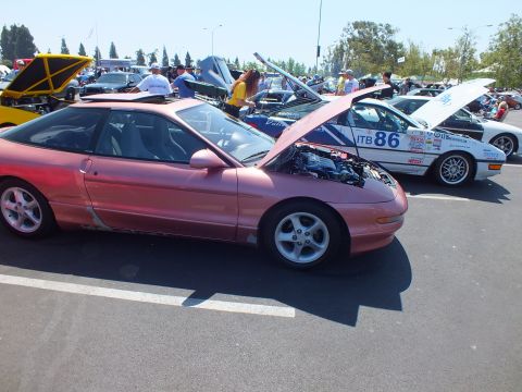 2003 Ford Crown Victoria - salmon-pink sports coupe with raised hood parked beside a white number-86 race car at a car show.